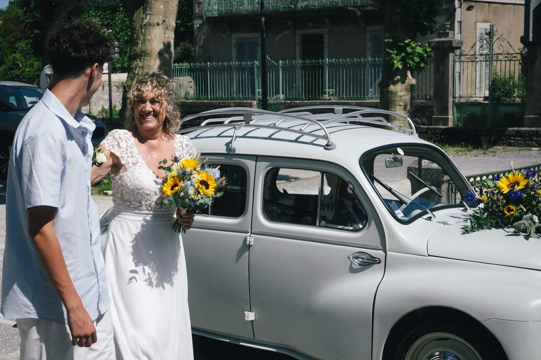 Mariage à Saint-Alban-Auriolles : la mariée près de la voiture ancienne, bouquet de tournesols à la main.