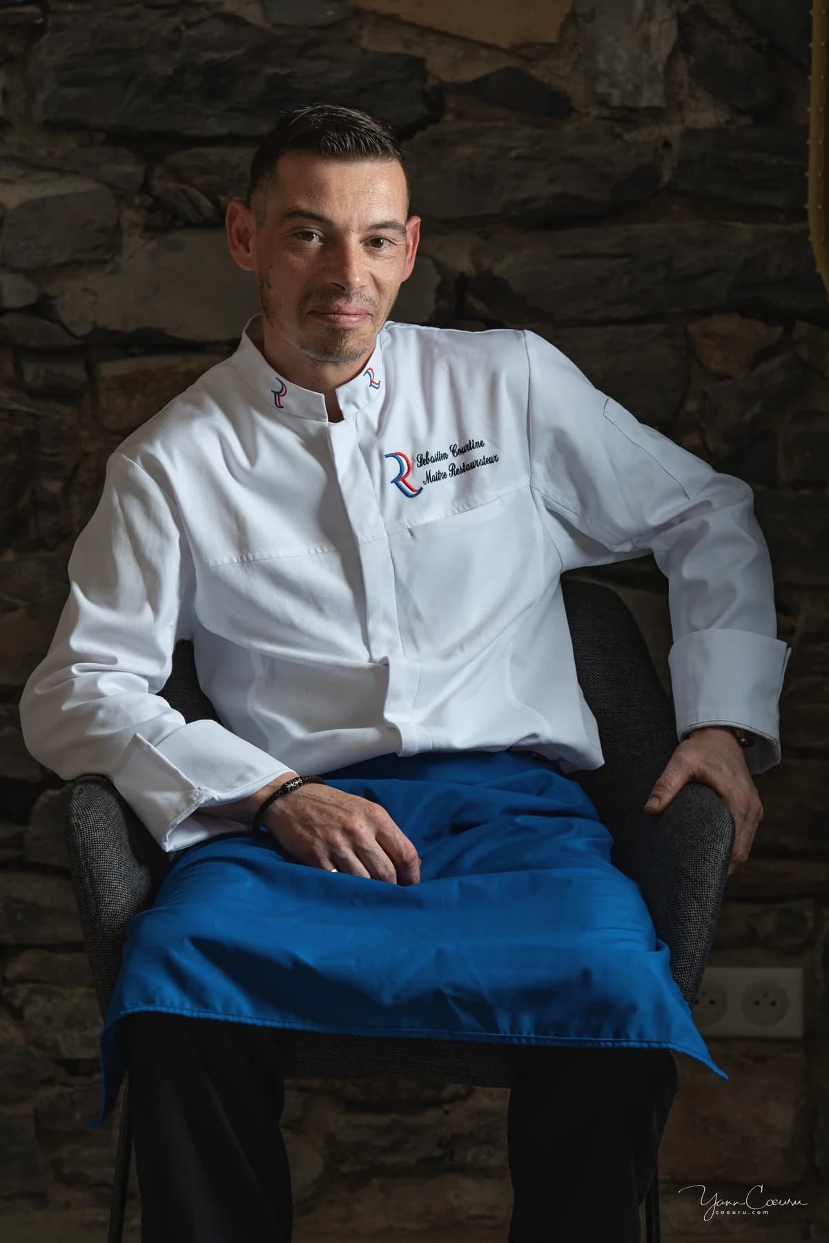 Portrait professionnel d'un chef restaurateur assis, veste blanche et tablier, studio mobile en Ardèche.