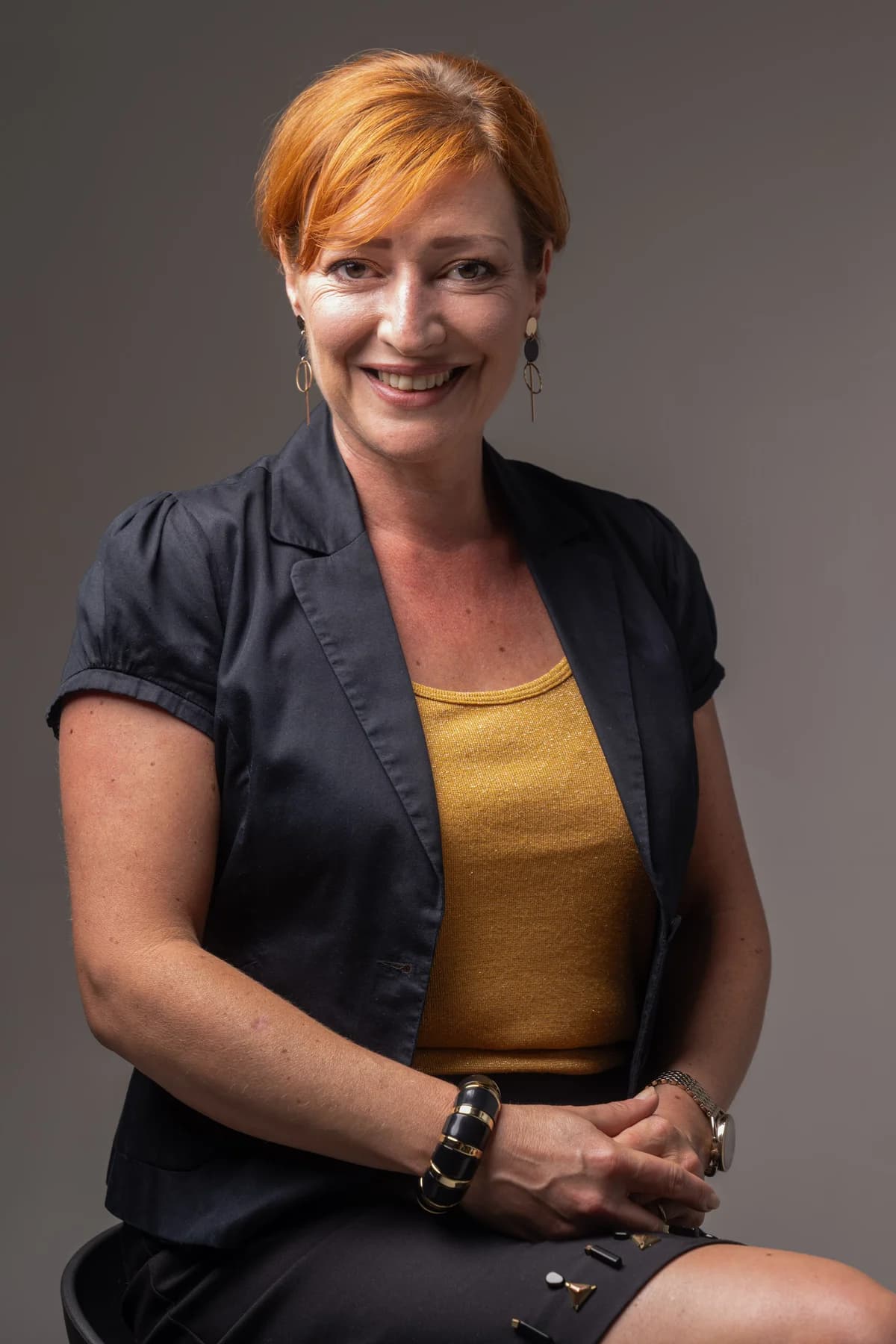 Portrait professionnel en studio — femme rousse souriante en veste noire, fond gris