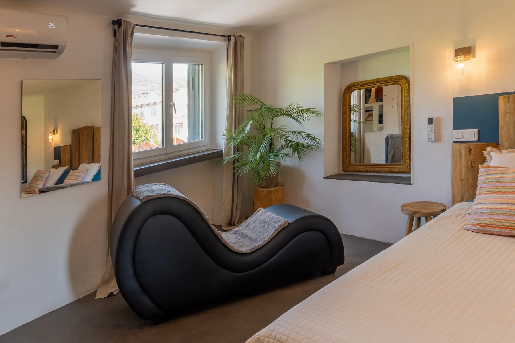 Immobilier Vaucluse : chambre lumineuse avec chaise lounge noire, miroir doré et palmier d'intérieur, ambiance cosy et moderne.