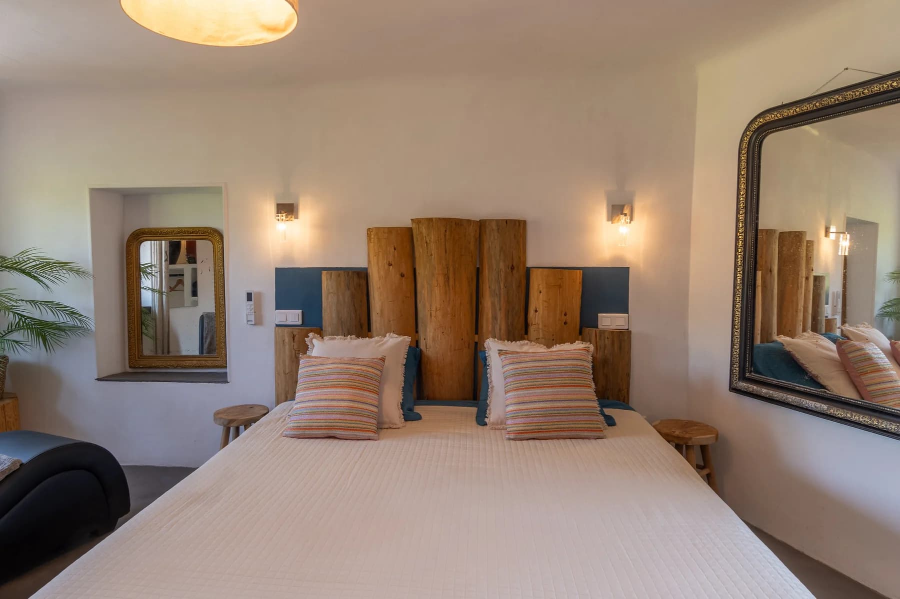 Immobilier Vaucluse : chambre lumineuse avec tête de lit en bois brut, coussins rayés et miroirs dorés vintage.