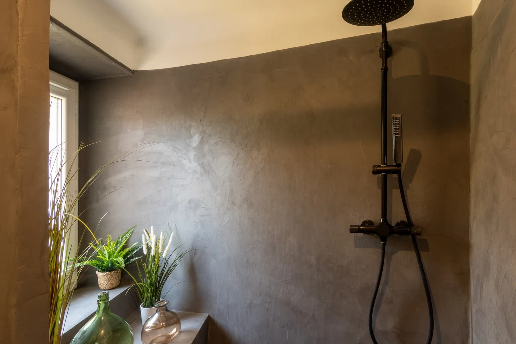 Immobilier Vaucluse – salle de bain en béton ciré avec douche à effet pluie noire et plantes vertes en lumière naturelle.