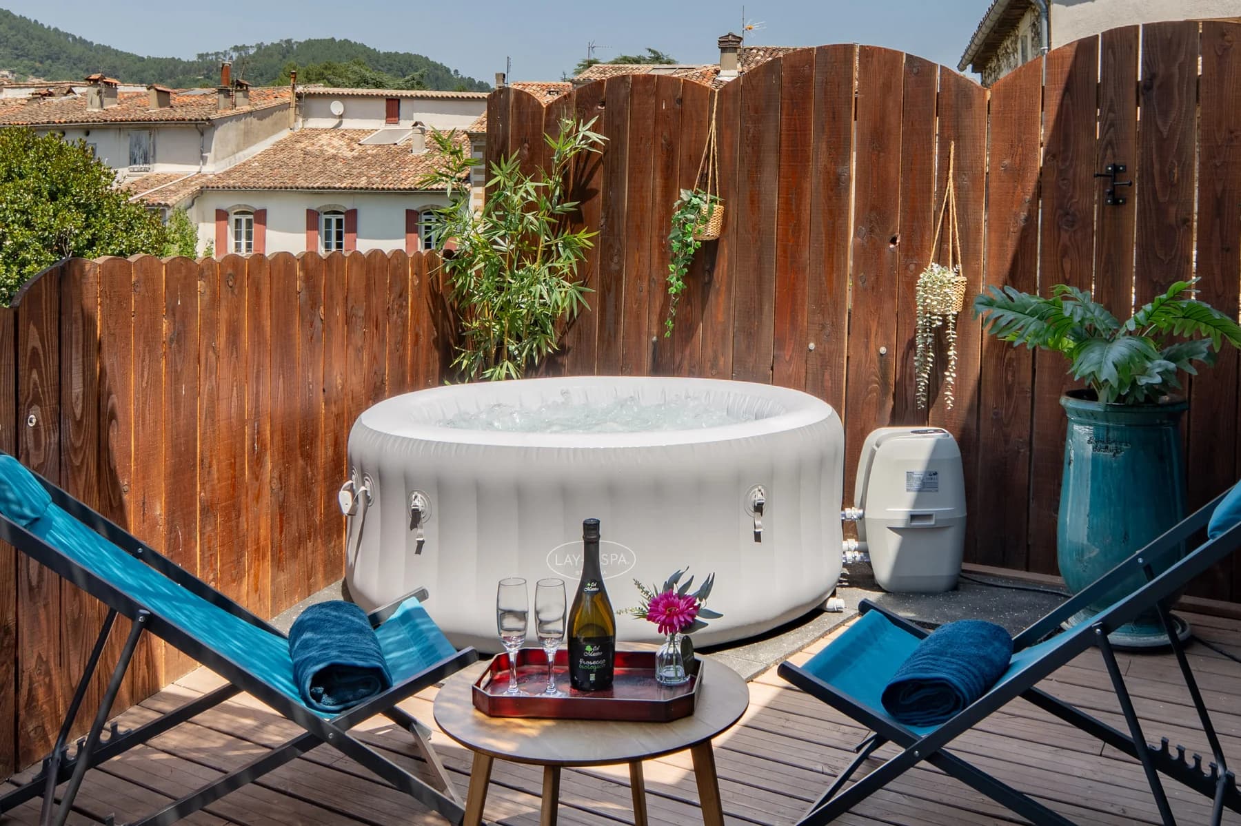 Immobilier Vaucluse, terrasse en bois avec spa gonflable, chaises longues bleues et plateau champagne au soleil.