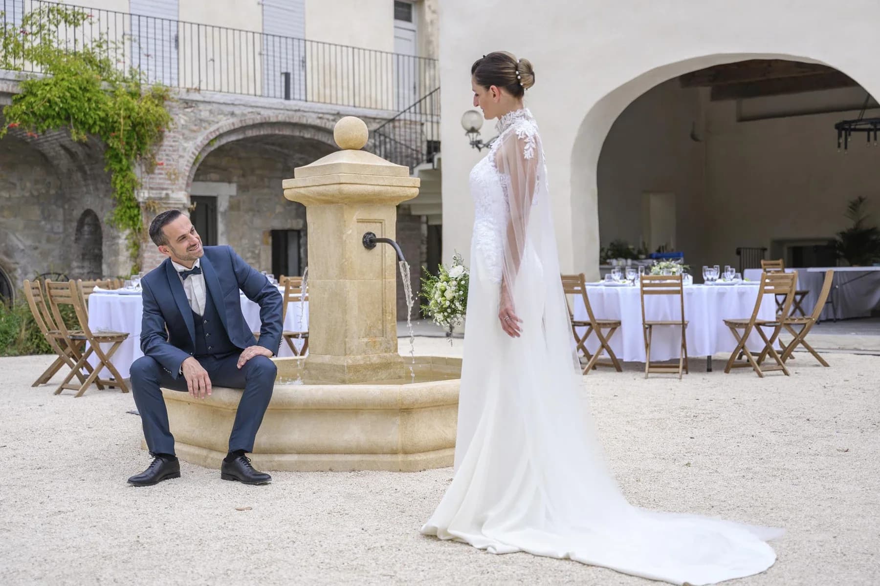 Mariage au Domaine de la Bedosse, à Alès : mariée en robe blanche et marié en costume bleu devant fontaine en pierre, cour méditerranéenne