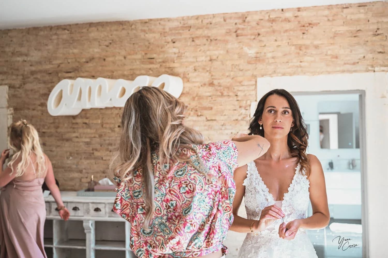 Mariages en intérieur, habillage de la mariée en robe dentelle blanche aidée par une amie, ambiance lumineuse et complice.