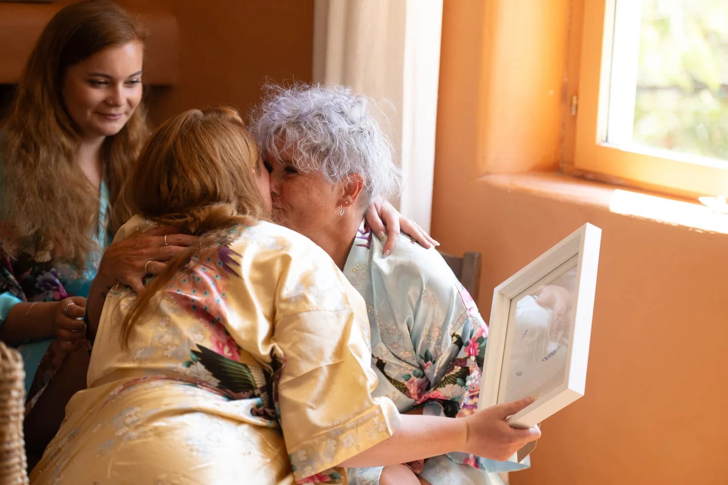 Mariages émouvant : accolade tendre entre deux femmes en robes de soie, lumière dorée et regard complice en arrière-plan.