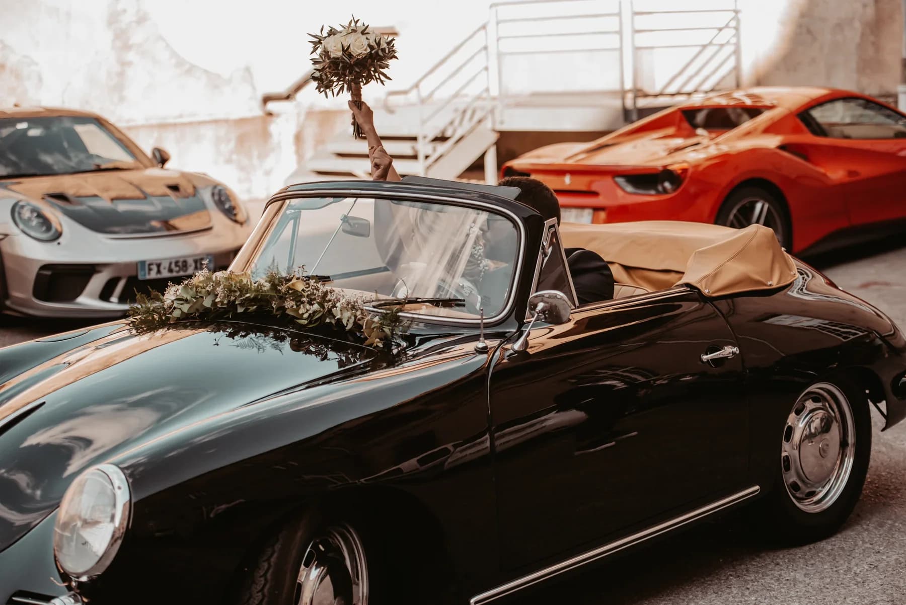 Mariages à Aubenas, mariée en cabriolet vintage noir brandissant son bouquet blanc entouré de voitures de sport.
