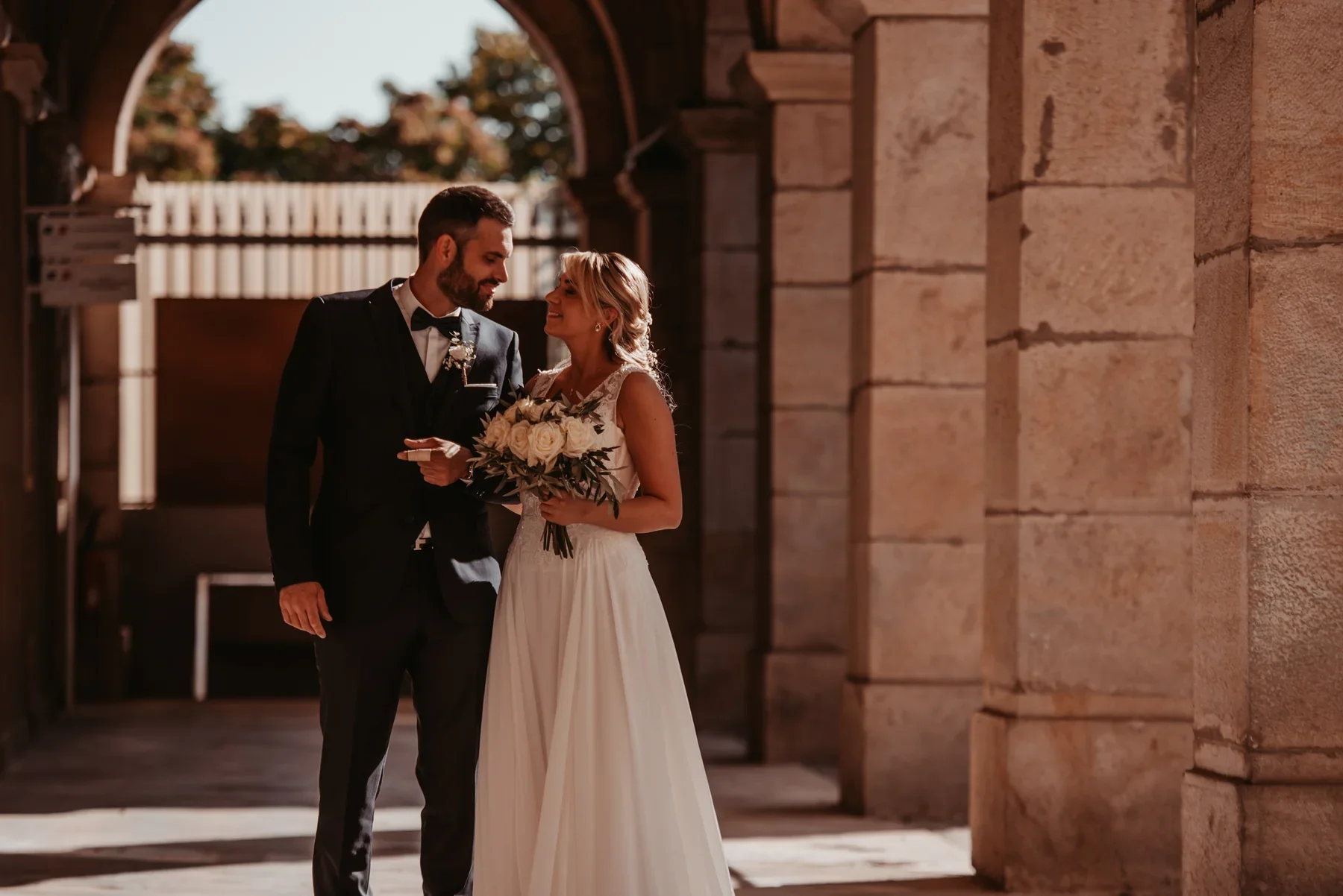 Ambre et Rémi sous les arcades en pierre dorée d'Aubenas, bouquet de roses blanches — mariage en Ardèche