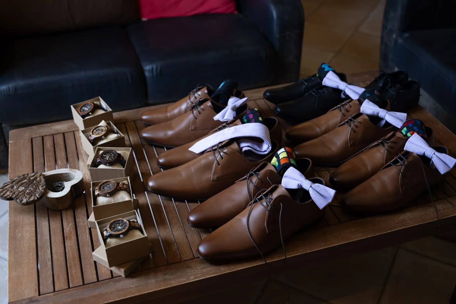 Mariages : détail des accessoires garçons d'honneur, chaussures marron, nœuds papillon et montres en bois disposés sur une table.