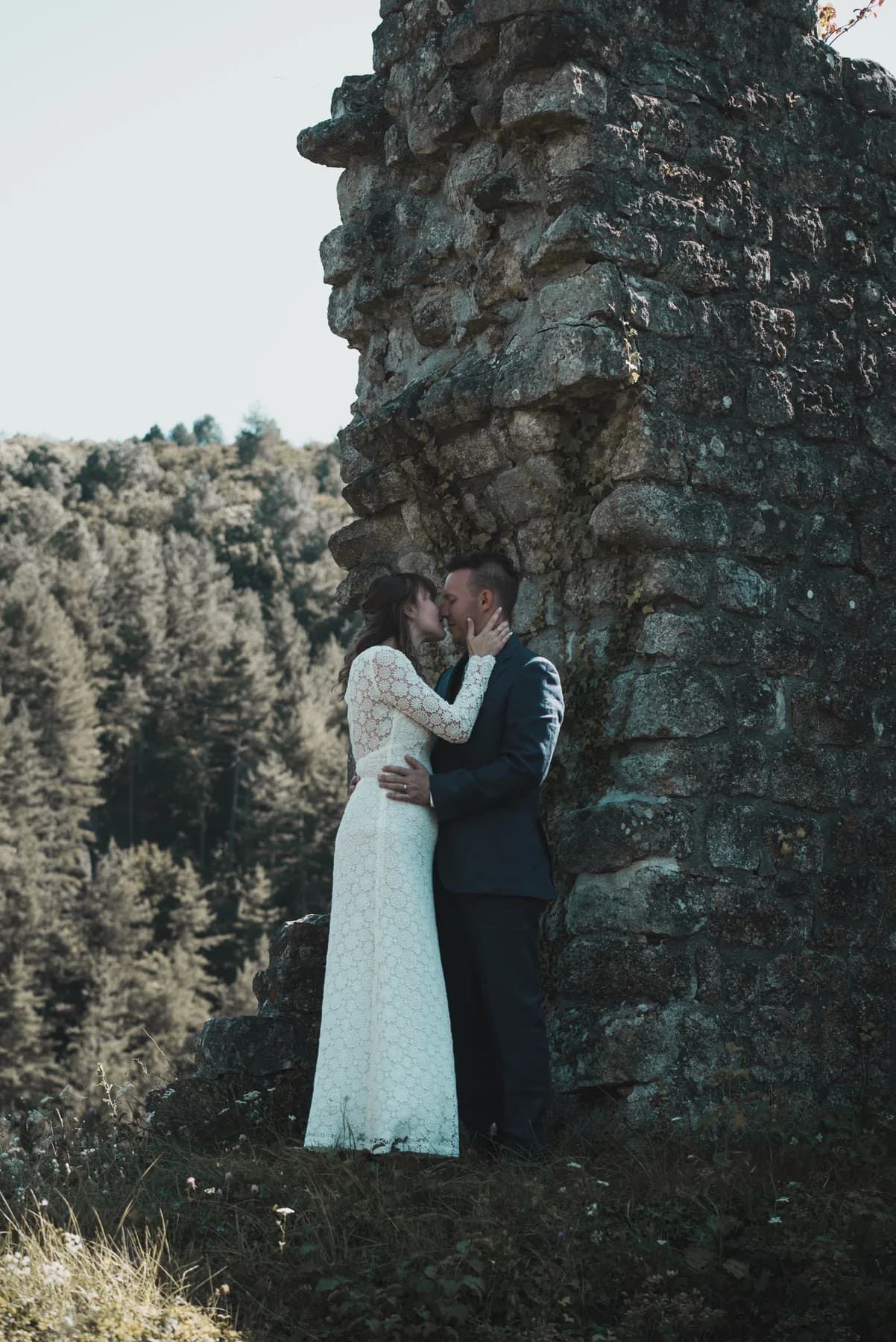 Mariages : couple s'embrassant en robe de dentelle blanche contre des ruines en pierre, forêt en arrière-plan.
