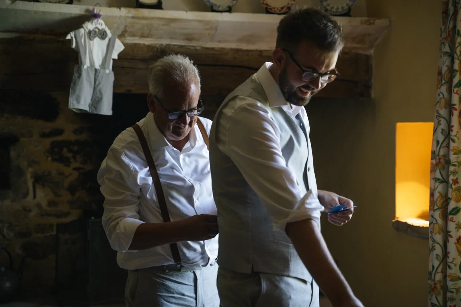 Mariages en intérieur rustique, deux hommes en costume gris se préparent dans une ambiance chaleureuse et complice.
