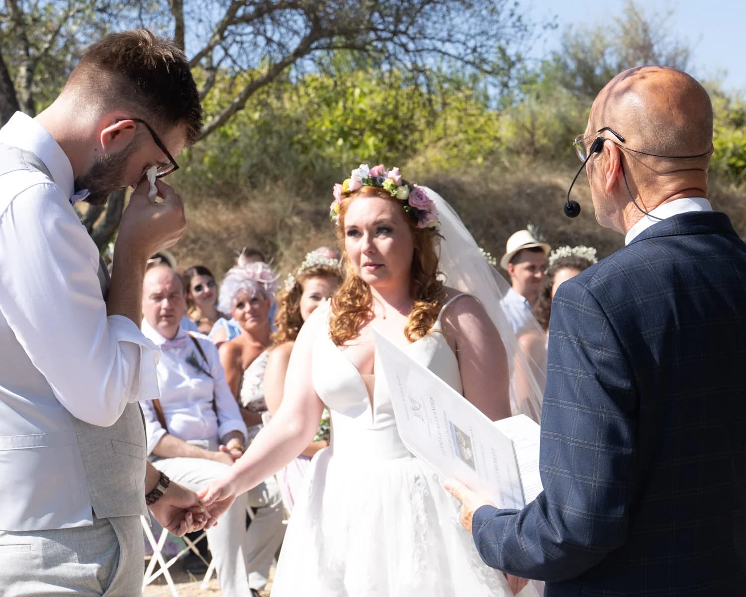 Mariages émouvant : marié en larmes tenant la main de sa mariée couronnée de fleurs lors d'une cérémonie en plein air.