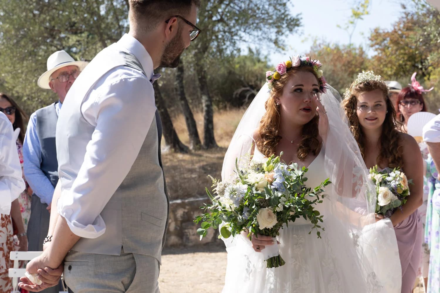Mariages : mariée émue en robe blanche et couronne florale face au marié lors d'une cérémonie en plein air.